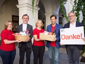 Mitarbeiter*innen der Caritas stehen in einem Innenhof und halten zwei mit Lebensmitteln gefüllte Körbe sowie ein Schild mit der Aufschrift "Danke!" in ihren Händen.