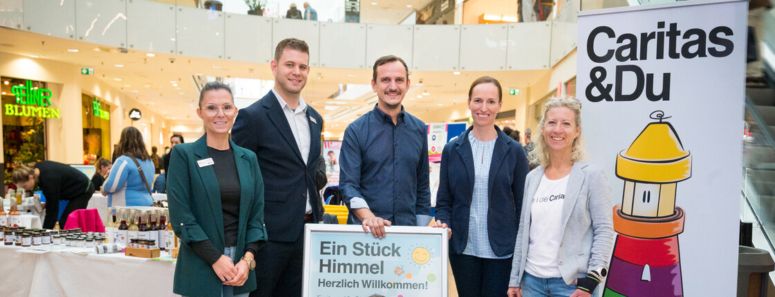 Mitarbeiter*innen der Caritas und drei kleine Mädchen stehen vor einem Marktstand in einem Einkaufzentrum und präsentieren mit Plakaten und Bannern die "Schule am Himmel".