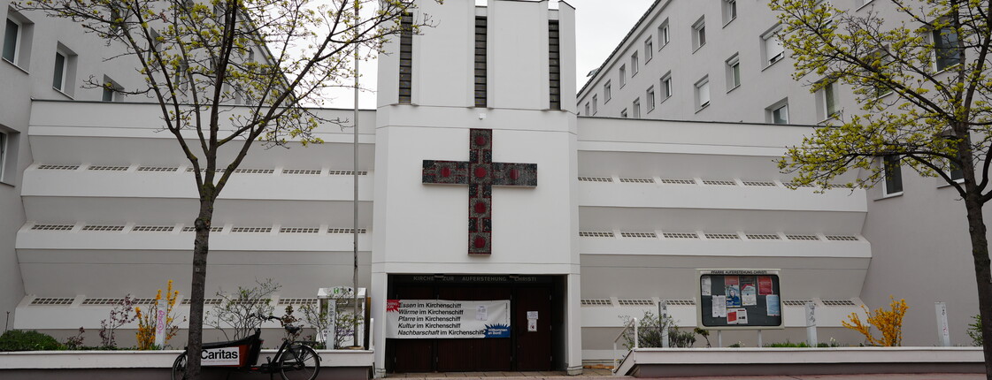 Das neue Le+O Kirchenschiff: Moderne weiße Kirchenfassade mit markantem dunklem Mosaikkreuz zwischen zwei Wohngebäuden.