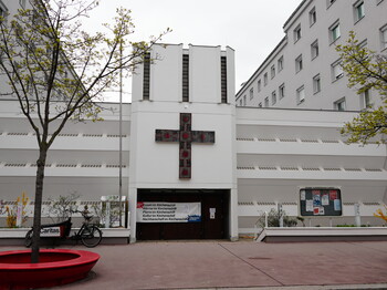 Das neue Le+O Kirchenschiff: Moderne weiße Kirchenfassade mit markantem dunklem Mosaikkreuz zwischen zwei Wohngebäuden.