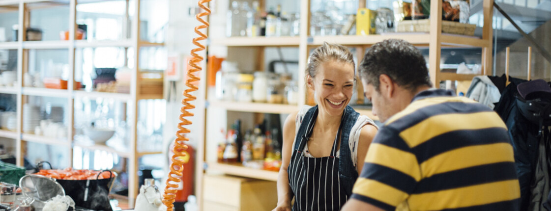 Zwei Personen bereiten gemeinsam frisches Gemüse in einer offenen Gemeinschaftsküche zu. Beide tragen Schürzen, schneiden Zutaten und lächeln einander an. Im Hintergrund sind Regale mit Kochutensilien und Lebensmitteln zu sehen. 