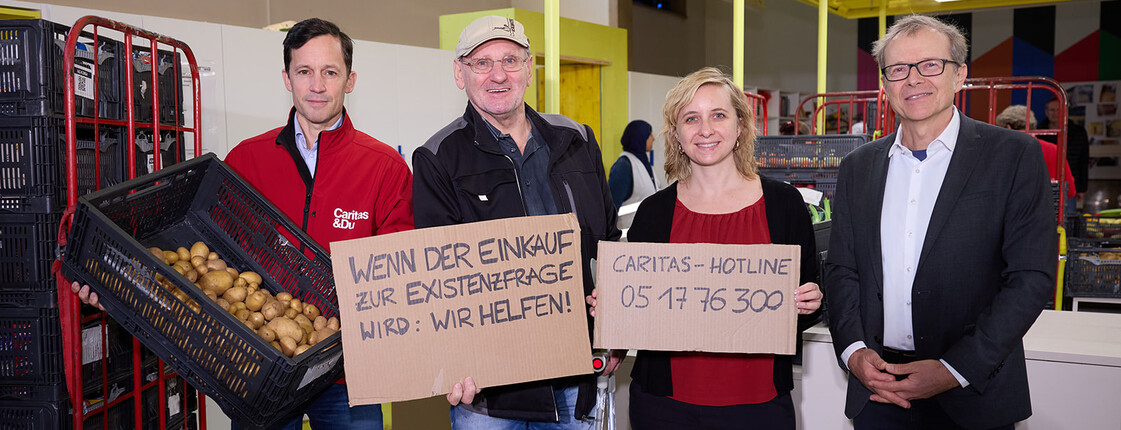 Vier Personen stehen in einer Lebensmittelausgabestelle der Caritas. Eine Person hält eine Kiste mit Kartoffeln, zwei andere halten Kartonschilder mit der Aufschrift „Wenn der Einkauf zur Existenzfrage wird: Wir helfen!“ und „Caritas-Hotline 05 17 76 300“. Im Hintergrund sind Regale und Lebensmittel zu sehen. Die Szene vermittelt Solidarität und Hilfe für Menschen in Not.