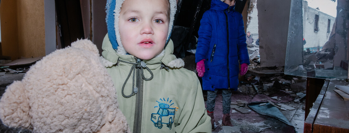 Ein kleiner Junge in Winterjacke und mit Haube hält einen Teddybären im Arm. Im Hintergrund sind die völlig zerstörten Möbel und Wände einer Wohnung zu sehen sowie auch ein traurig dreinschauendes kleines Mädchen.