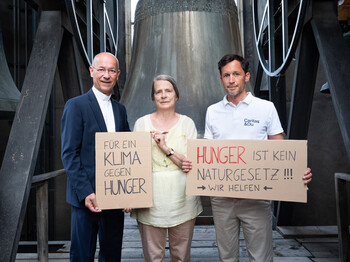 Die Meteorologin Helga Kromp-Kolb und die Direktoren der Caritas halten vor einer Kirchenglocke Kartonschilder mit der Aufschrift "Für ein Klima gegen Hunger" und "Hunger ist kein Naturgesetz! Wir helfen".