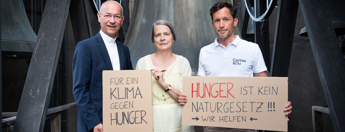 Die Meteorologin Helga Kromp-Kolb und die Direktoren der Caritas halten vor einer Kirchenglocke Kartonschilder mit der Aufschrift "Für ein Klima gegen Hunger" und "Hunger ist kein Naturgesetz! Wir helfen".