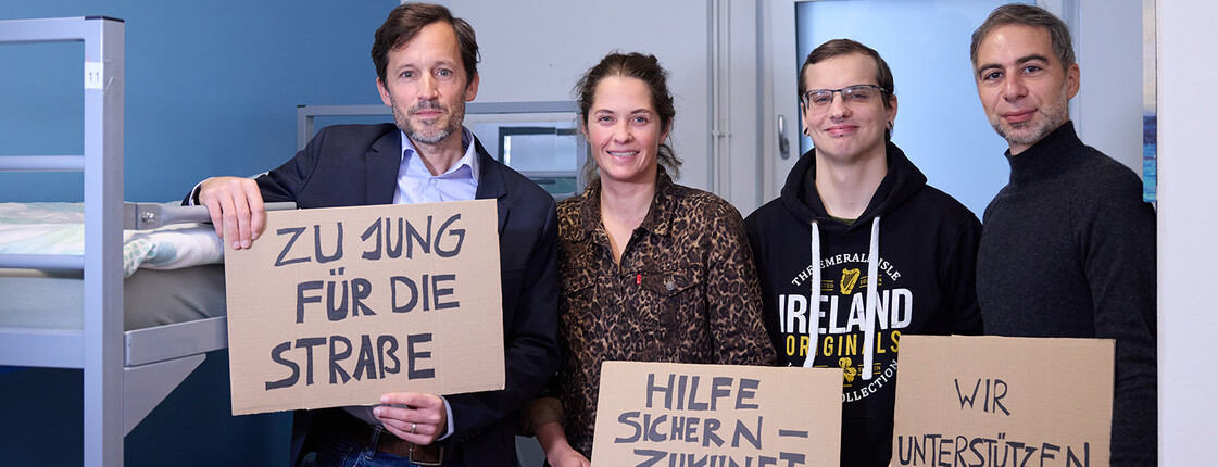 Vier Personen stehen in einem Schlafraum mit Etagenbetten und halten Schilder aus Karton mit handgeschriebenen Botschaften: „Zu jung für die Straße“, „Hilfe sichern – Zukunft schenken“ und „Wir unterstützen“. Es handelt sich um einen Pressetermin der Caritas zur Kampagne für junge Wohnungslose. Die Personen auf dem Foto sind Klaus Schwertner, Tom Adrian (Leiter von a_way), Maresi Kienzer (Leiterin Wohnhaus JUCA) und eine weitere Person.